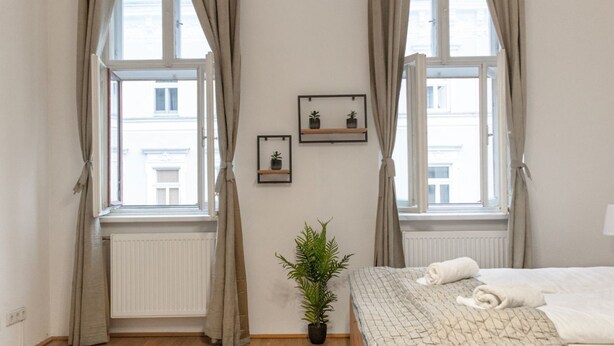 Une chambre à coucher avec un lit, deux fenêtres avec des rideaux, un radiateur, une plante en pot et deux tableaux encadrés accrochés au mur.