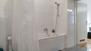 Une salle de bain avec des murs carrelés de blanc, une douche équipée d’un pommeau de douche amovible et un porte-rouleau de papier toilette visible.