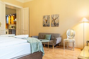 A hotel room with a bed, a sofa, a chair, a lamp, and two wall-mounted paintings.