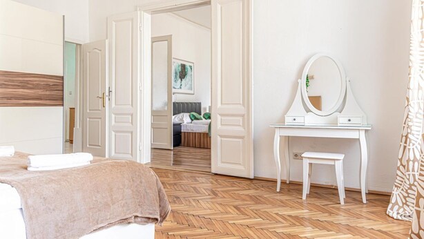 Ein Hotelzimmer mit einem Bett, einem Schminktisch mit Spiegel, einer Bank und einer offenen Tür, die in einen anderen Raum führt.