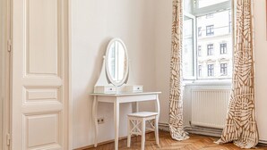 Ein Zimmer mit weißem Schminktisch, einem Spiegel, einem kleinen Hocker, einem Fenster mit gemusterten Vorhängen und Blick auf Gebäude.
