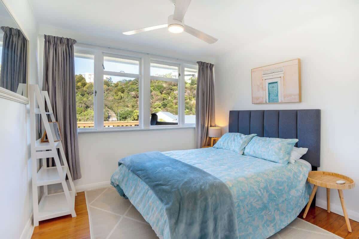 A bedroom with a bed, a ceiling fan, a window with curtains, a framed picture on the wall, and a small side table.