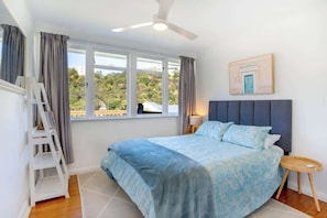 Un dormitorio con cama, ventilador de techo, ventana con cortinas, un cuadro en la pared y una mesita de noche.