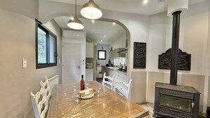Un comedor con una mesa de madera, sillas blancas, iluminación colgante, un estufa de leña negra y una cocina al fondo.