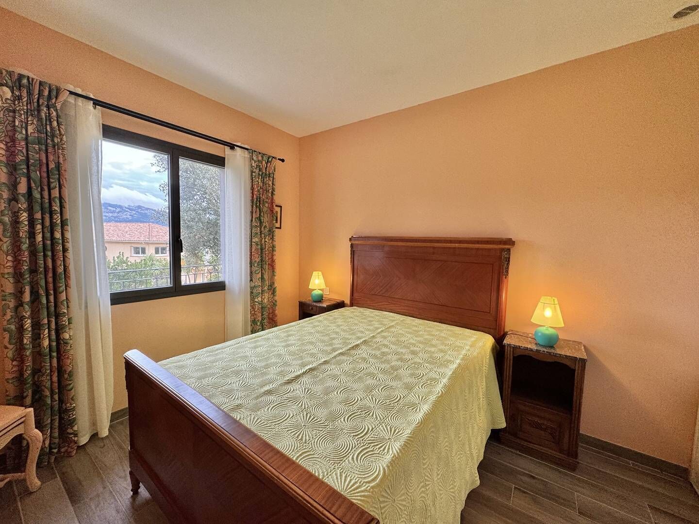 Ein Schlafzimmer mit einem hölzernen Bett, zwei Nachttischlampen, einem Fenster mit Vorhängen und Blick auf Gebäude.