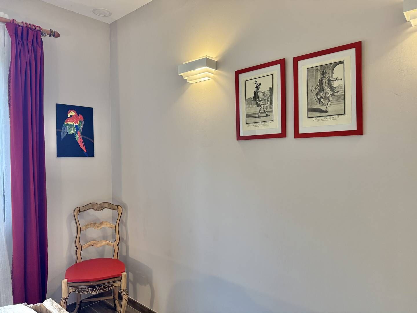 A room with a wooden chair with a red cushion, a framed black and white artwork, and a wall-mounted light fixture.