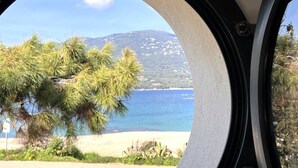 Une vue à travers une fenêtre circulaire, avec une plage, un arbre et une montagne en arrière-plan.