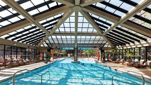 Una piscina interior con techo alto y paneles de vidrio, rodeada de zonas de descanso con sillones.