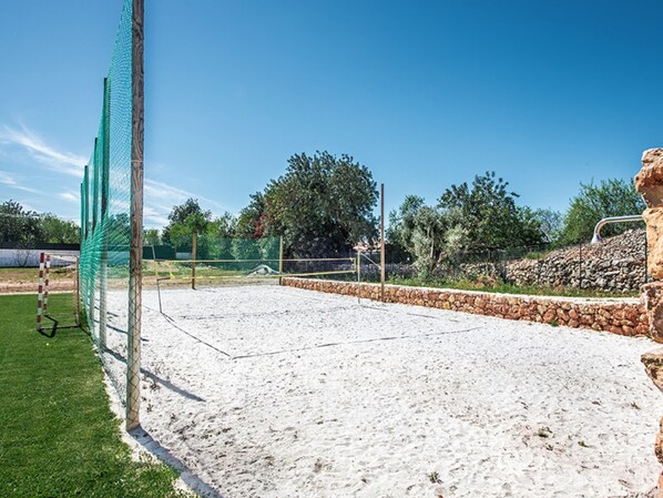 Un terrain de beach-volley sablonneux, avec un filet, entouré d’un mur de pierres et de verdure.