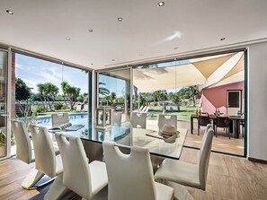 Un espace repas moderne avec une table en verre et des chaises blanches, donnant sur une piscine et un coin salon extérieur.
