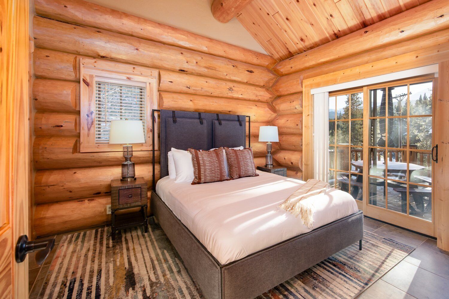 Ein Schlafzimmer in einer Blockhütte mit einem Bett, zwei Nachttischen, einem Fenster und Blick ins Freie.