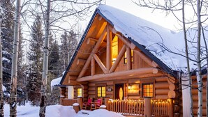 Lumihunnun peittämä hirsimökki, jonka ympärillä on puita.