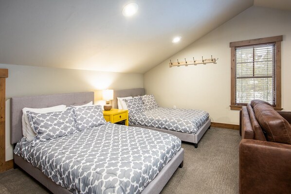 A bedroom with two beds, a nightstand, a window with blinds, and a leather bench.
