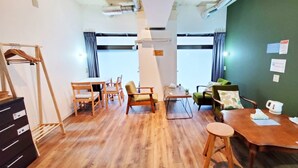 A room with a wooden floor, a dining table with chairs, a sofa, a wooden wardrobe, and a green wall. - EZO Run Sapporo - Hostel (Sapporo)