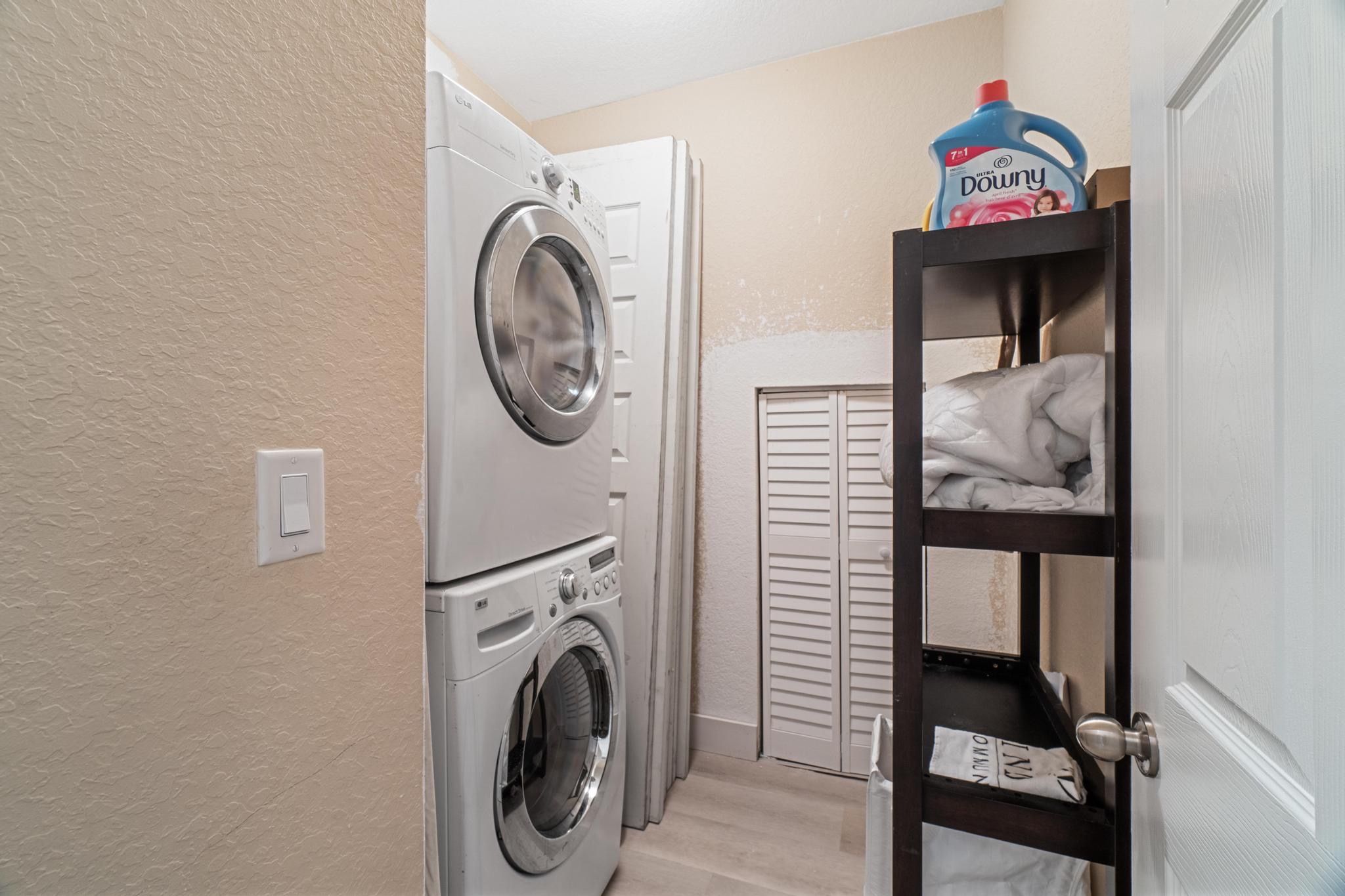 A laundry room with a front-loading washer and dryer, a shelf with a detergent bottle, and a door with a towel rack.