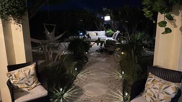 En udendørs patio med sofa og stole, omgivet af planter og en stensti.