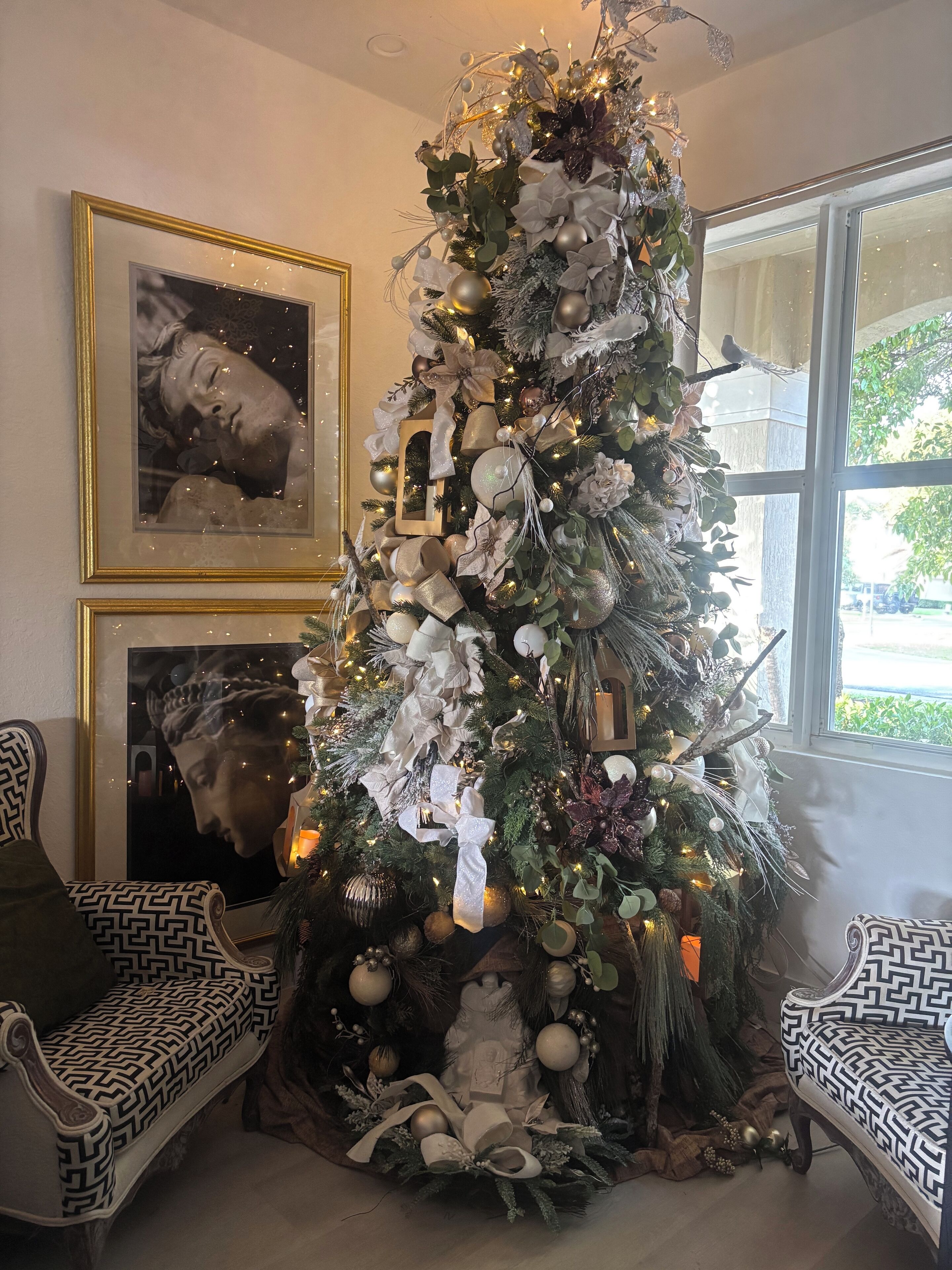 A Christmas tree decorated with white and gold ornaments, ribbons, and lights, placed in a room with patterned chairs and framed artwork on the walls.
