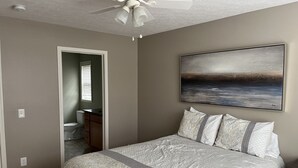 Un dormitorio con cama, ventilador de techo, un cuadro y un baño visible a través de una puerta abierta.