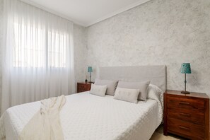 Une chambre à coucher avec un lit, deux tables de chevet, une lampe et une fenêtre avec des rideaux légers.