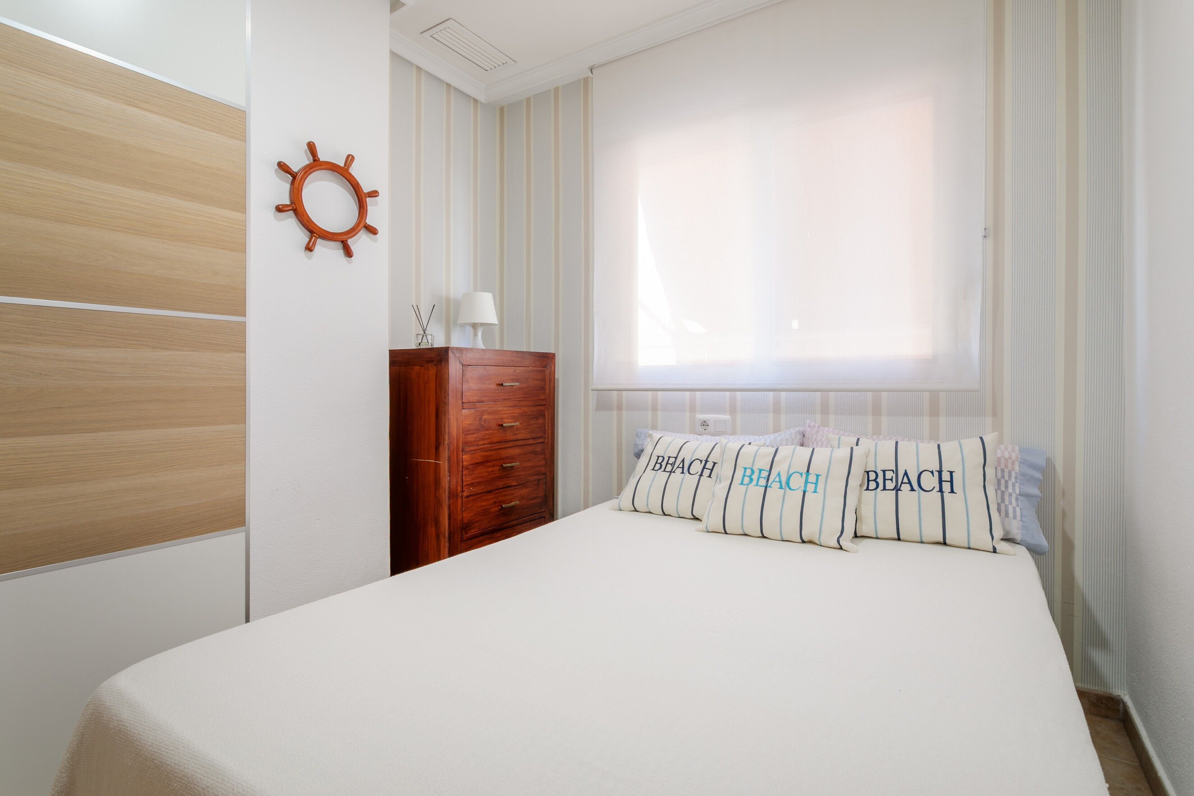 A bedroom with a bed, a wooden dresser, a wall-mounted sun decoration, and striped curtains.