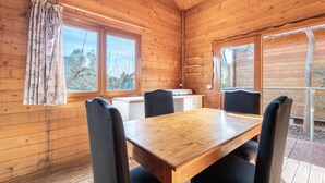 A wooden cabin interior with a dining table, chairs, a window with curtains, and a view of trees outside. - Holiday Home 'Barraca Adaptado B' with Mountain View, Wi-Fi and Air Conditioning (Bárig)