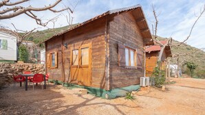 A wooden cabin with a stone wall, a table with chairs, and a dirt path. - Holiday Home 'Barraca' with Mountain View, Wi-Fi and Air Conditioning (Bárig)