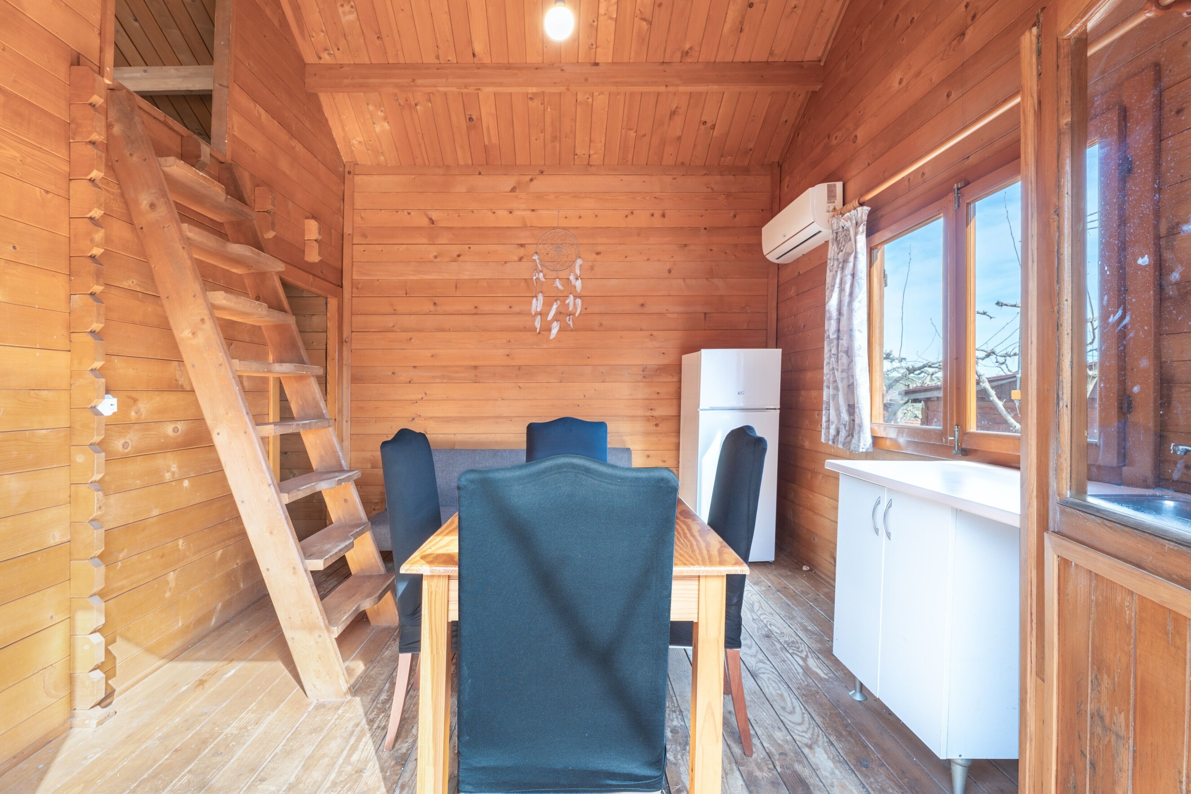 Interior de una cabaña de madera con una mesa de comedor, sillas, una escalera, un refrigerador y una ventana.