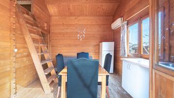 Interior de una cabaña de madera con una mesa de comedor, sillas, una escalera, un refrigerador y una ventana.