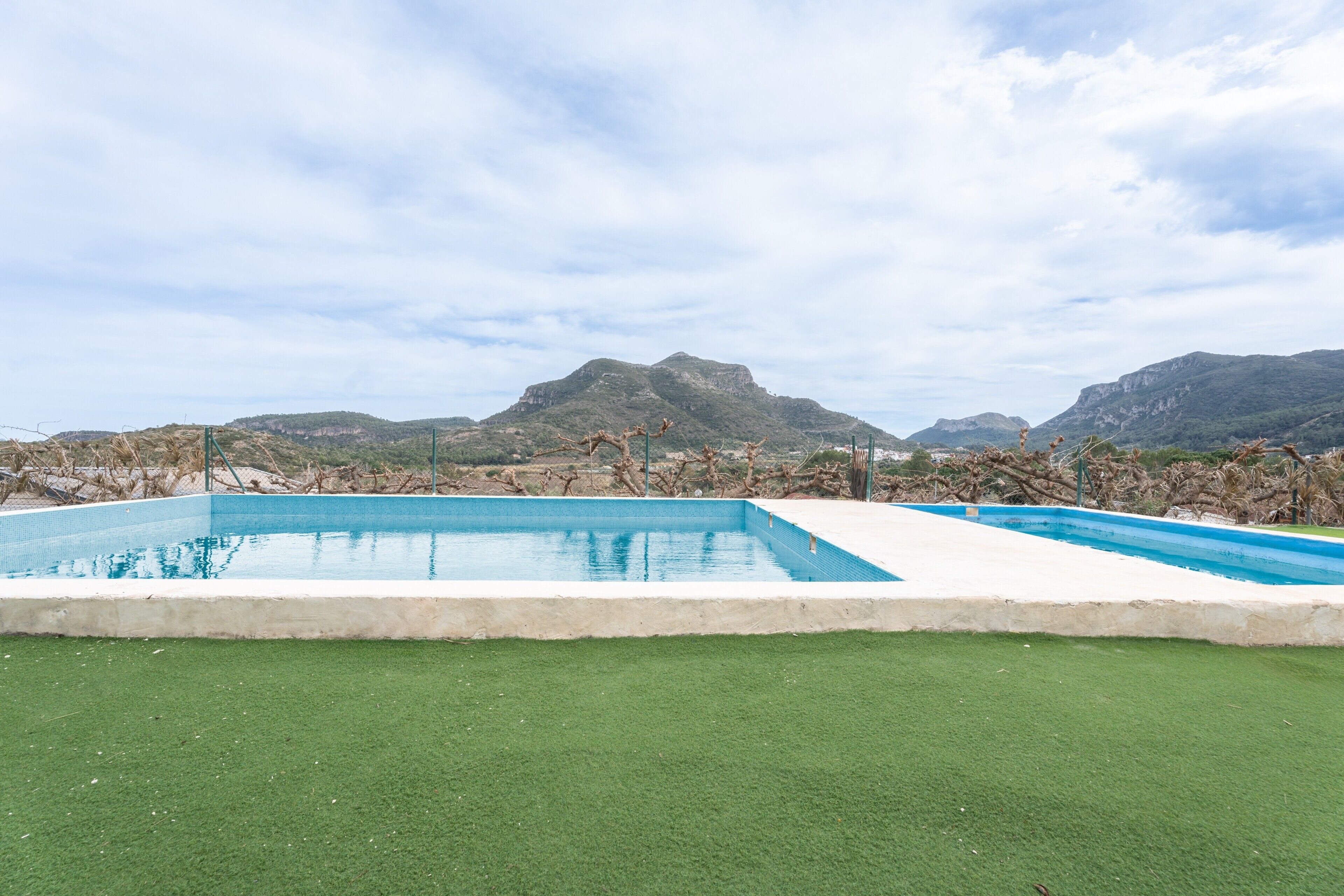 Piscina rectangular con borde blanco, rodeada de césped artificial verde, con montañas al fondo bajo un cielo parcialmente nublado.