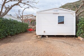 Una casa móvil blanca con ventana y puerta, ubicada en un camino de grava con una silla roja y un seto en primer plano.