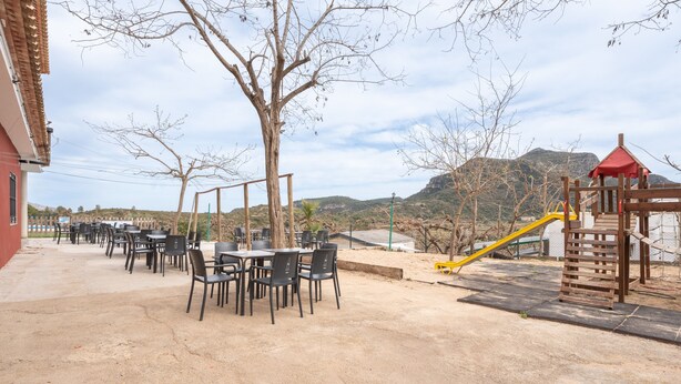 An outdoor patio with tables and chairs, a playground with a slide, and a view of hills in the background. - Holiday Home 'BW 2018 IV' with Mountain View, Wi-Fi and Air Conditioning (Bárig)