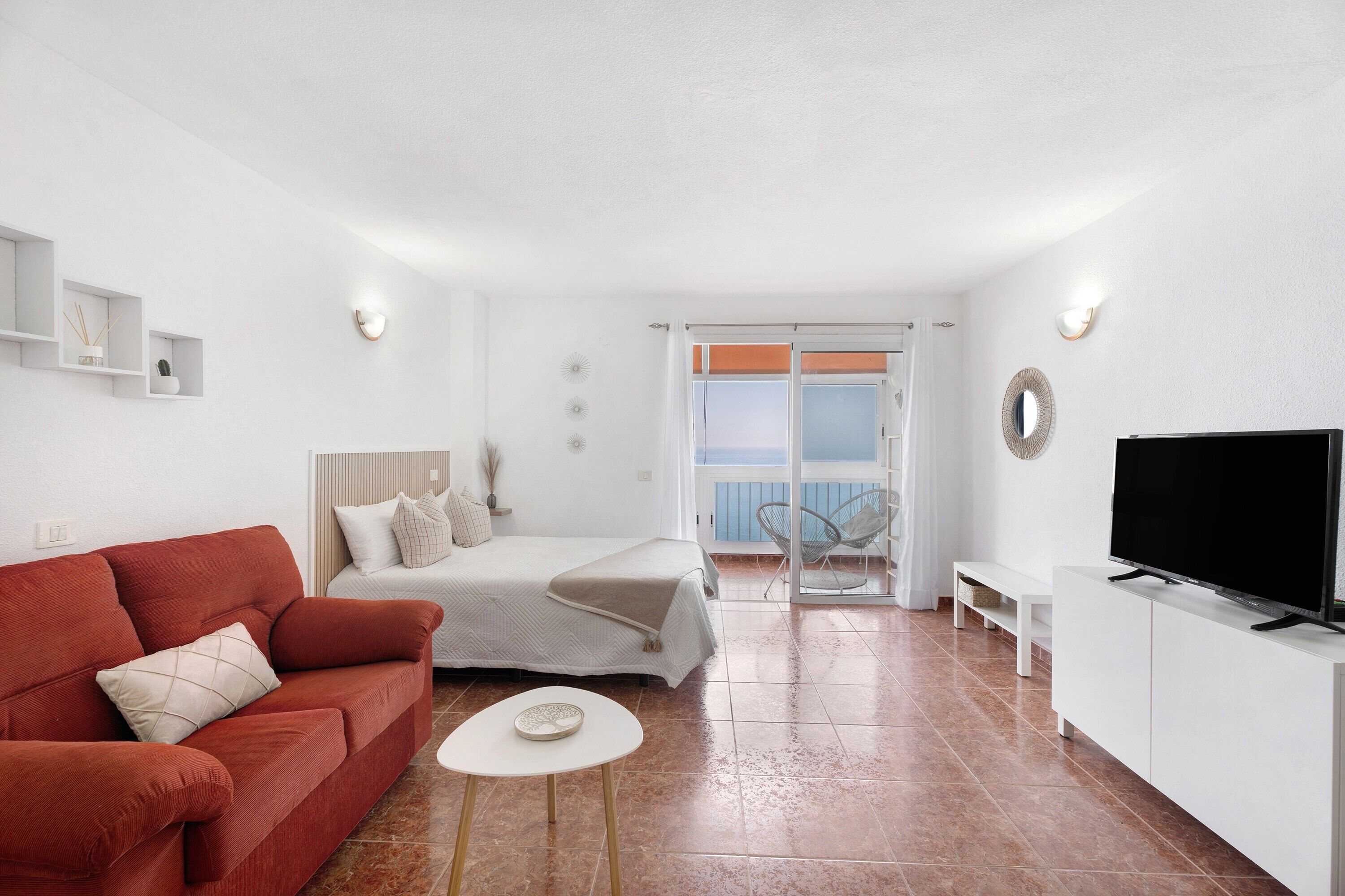 Una sala moderna con un sofá rojo, una mesa de centro blanca, una cama con cabecera blanca, un televisor sobre un mueble blanco y un balcón con una silla.