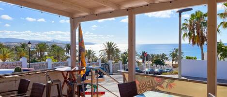 A covered outdoor seating area with wicker chairs and a table, overlooking a beach and palm trees.