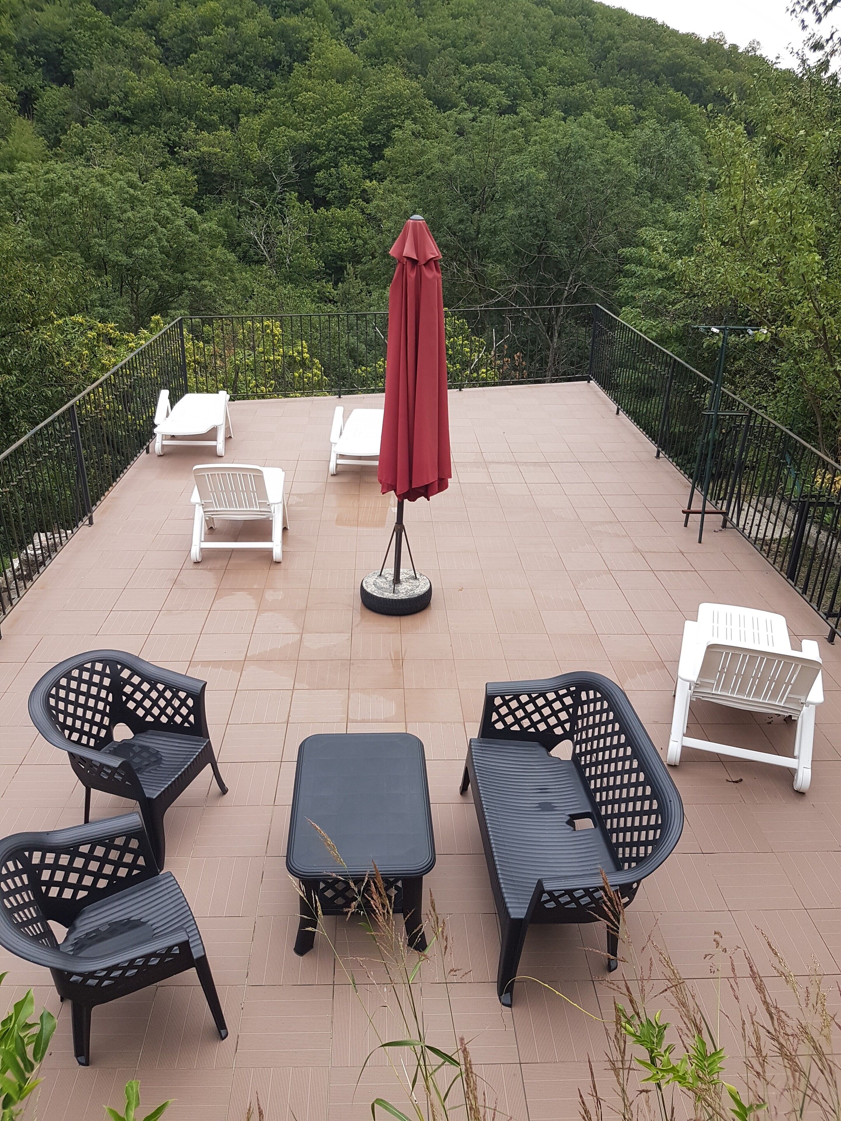 Une terrasse aménagée avec du mobilier d’extérieur, un parasol rouge et une vue sur une colline boisée.