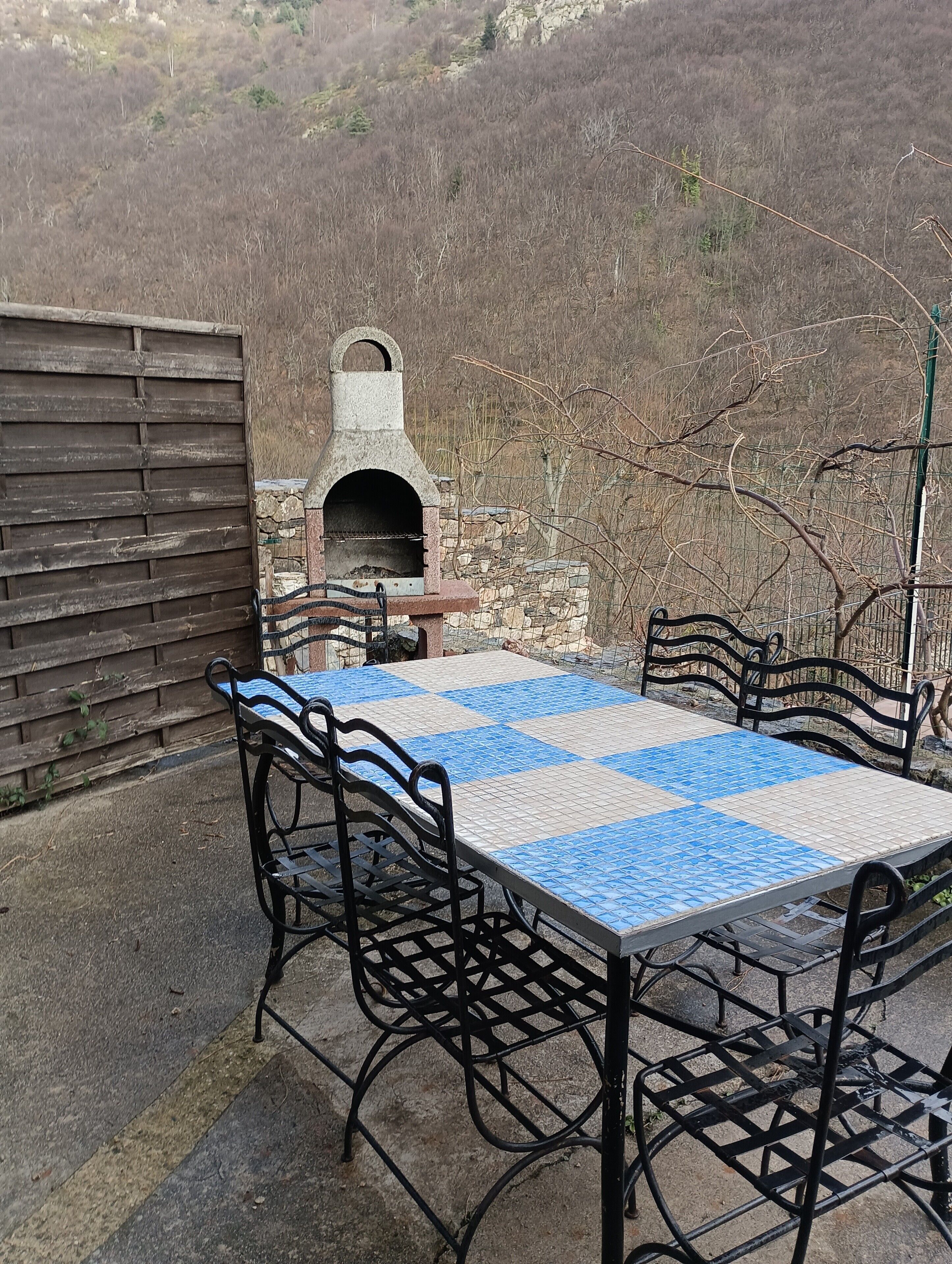 Una veranda esterna con un tavolo a scacchi, sedie in metallo e un forno in pietra.