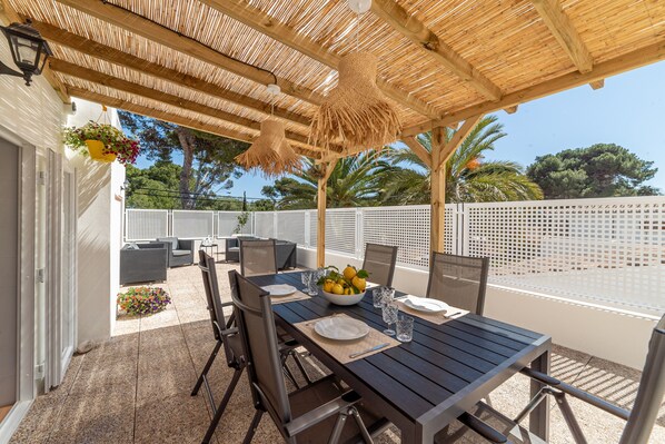 Une terrasse extérieure avec une table en bois dressée pour le repas, entourée de chaises, et un toit de chaume au-dessus.