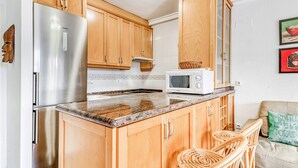 A kitchen with wooden cabinets, a stainless steel refrigerator, a microwave, and a wicker chair. - Apartment 'Casa Molins' with Shared Pool, Private Terrace and Wi-Fi (Dénia)