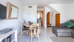 Un soggiorno con un divano grigio, un'area pranzo con tavolo e sedie, una TV su un mobile, e una scala che conduce a un'altra stanza.