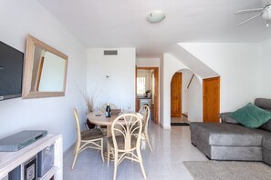 Un salon avec un canapé gris, un coin repas avec une table et des chaises, un téléviseur posé sur un meuble et un escalier menant à une autre pièce.