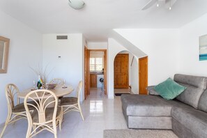 Un coin repas avec une table ronde et des chaises, une cuisine en arrière-plan et un canapé gris agrémenté d’un coussin vert.