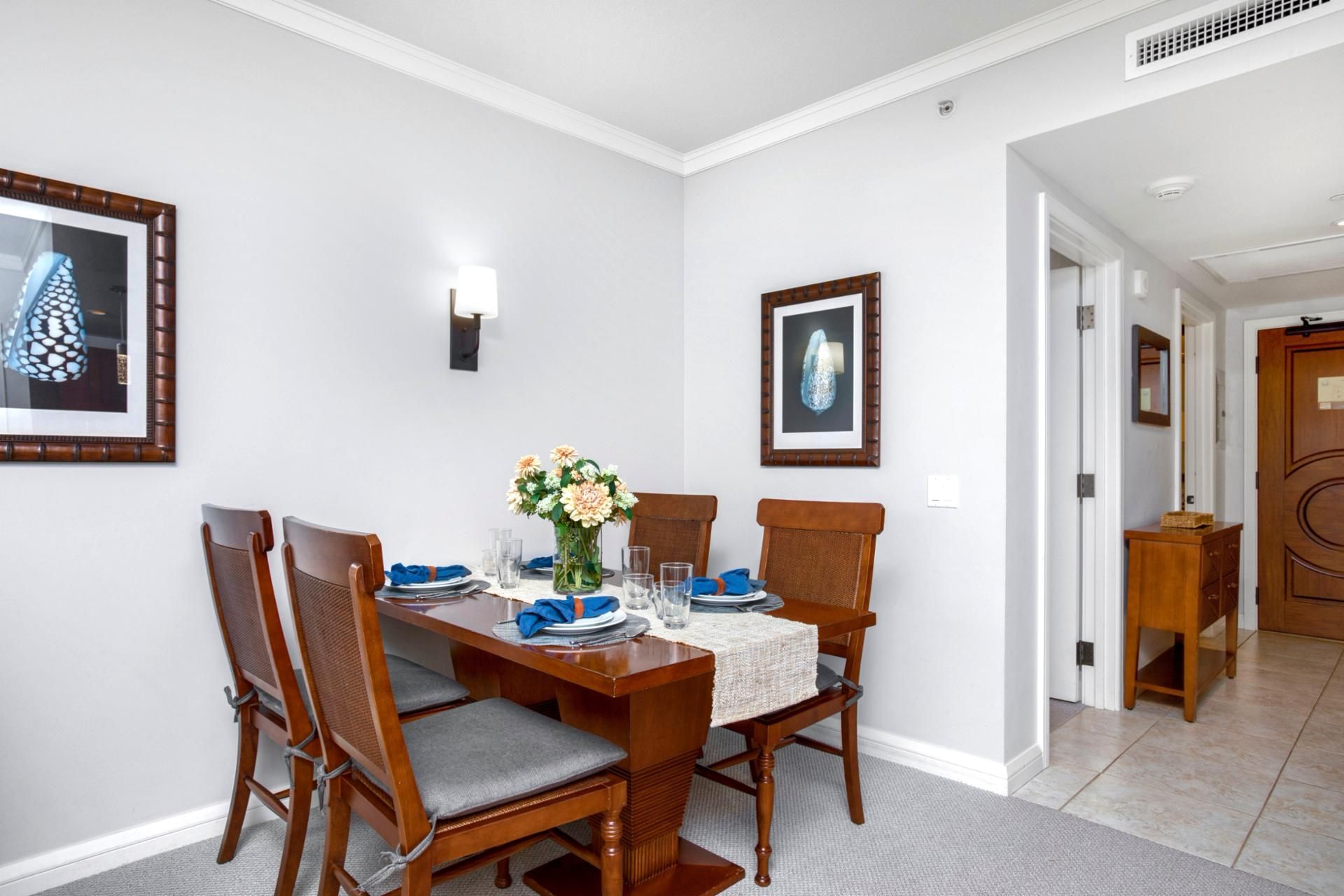A dining area with a wooden table set for four, surrounded by wicker chairs with grey cushions. There are framed pictures on the walls, a vase with flowers on the table, and a small wooden side table with a basket.