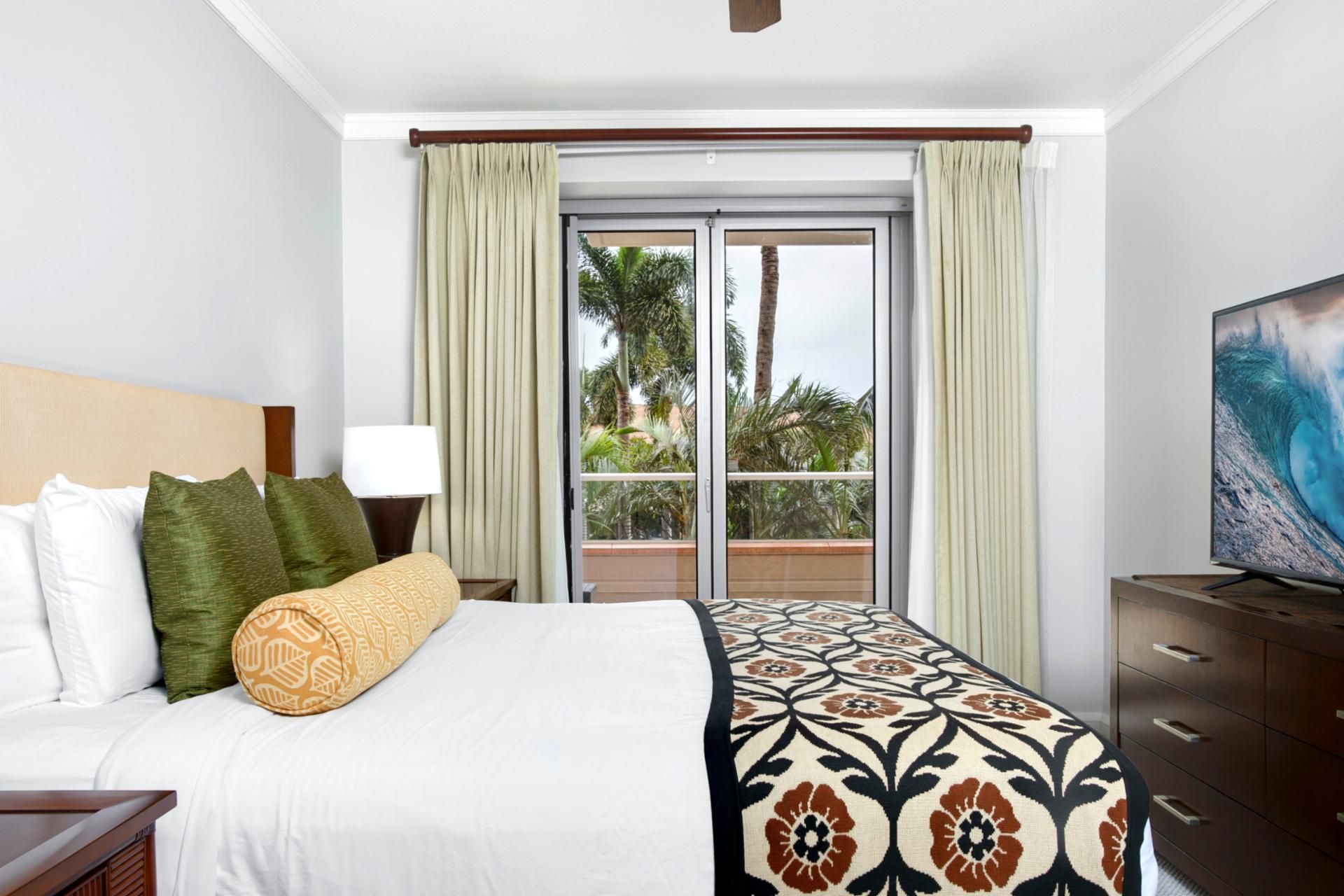 A hotel room with a bed, a nightstand, a TV, a window with curtains, and a view of palm trees.