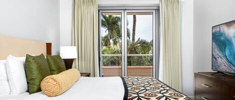 A hotel room with a bed, a nightstand, a TV, a window with curtains, and a view of palm trees.