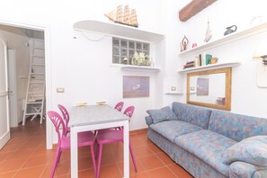 Une pièce avec une table blanche, des chaises roses, un canapé à motifs, une fenêtre décorée d’un motif de bateau, et une étagère contenant des livres et une maquette de bateau.