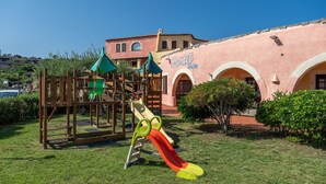 Ein Spielplatz mit Rutsche und Klettergerüst, ein Gebäude mit Bögen und einem Schild, auf dem 'Market' steht, sowie ein strahlend blauer Himmel.