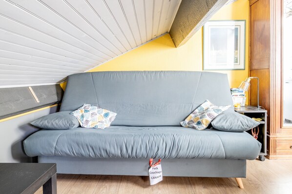 A grey sofa with patterned cushions, a small table, and a wall-mounted shelf with a lamp and a framed picture.