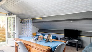 Una habitació amb un escriptori de fusta, dues cadires, un llit amb edredó blau, una televisió sobre un moble i una finestra amb cortines estampades.
