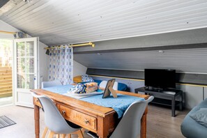 Une pièce avec un bureau en bois, deux chaises, un lit recouvert d’une courtepointe bleue, un téléviseur posé sur un meuble et une fenêtre ornée de rideaux à motifs.