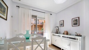Un comedor con una mesa de vidrio, sillas blancas, un armario blanco y una ventana con cortinas.