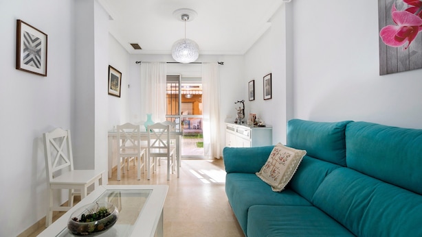 Una sala de estar con un sofá color esmeralda, una mesa de centro de vidrio y un comedor blanco con sillas.